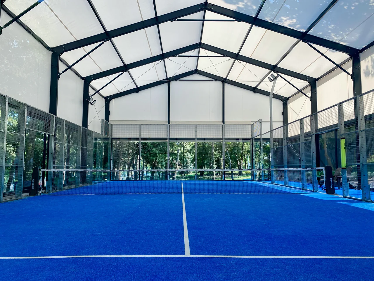 Blue padel court beneath a translucent roof, with a net, glass walls & trees outside